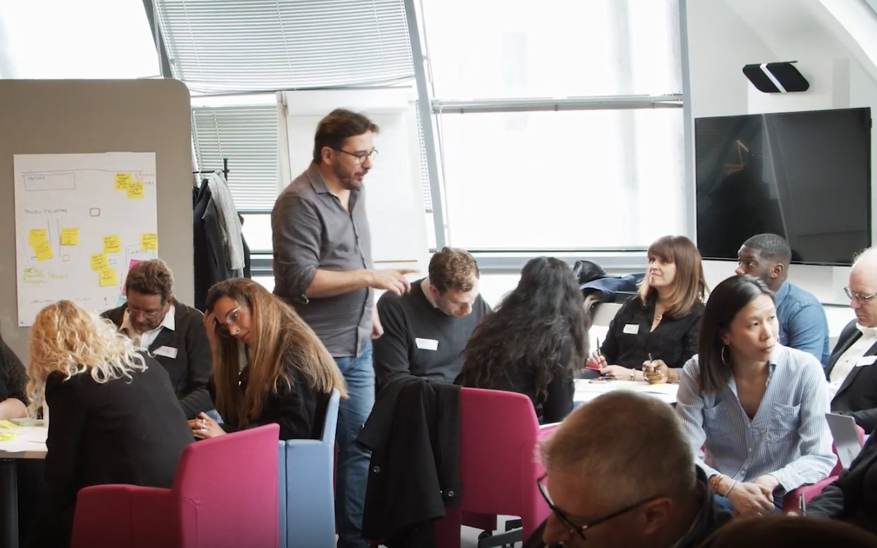 Atelier d'intelligence collective : une quinzaine de participants autour de tables de travail, un animateur debout devant un mur de post-its.